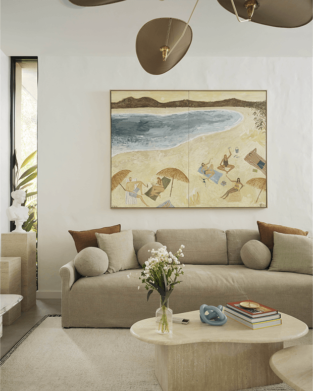 Warm, refined living room with a large upholstered neutral sofa, sculptural round coffee table, textured light carpet, and soft off-white walls. A coastal beach artwork hangs above the sofa, while sculptural ceiling fixtures, cushions, books, and a floral arrangement add decorative detail. A narrow glazed opening and small stair area appear at the left.