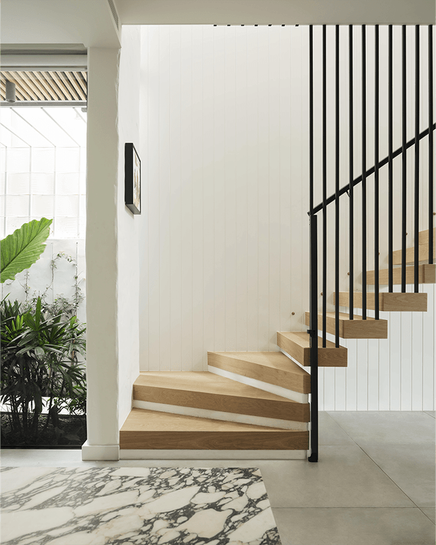 Bright contemporary stairway with floating light timber treads, slim black vertical metal balustrades, and white vertical wall panelling. The staircase is adjacent to a glazed indoor garden or sunroom, with pale concrete-look flooring and a bold marble-patterned floor section in the foreground.