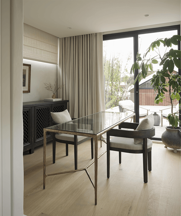 Refined dining area with a rectangular glass-top dining table and upholstered dark-framed chairs, set against pale timber flooring, full-height curtains, a dark decorative sideboard, and large black-framed glazing. A potted indoor tree and outdoor greenery visible through the windows add a natural element. The space has a calm, contemporary and softly neutral character.