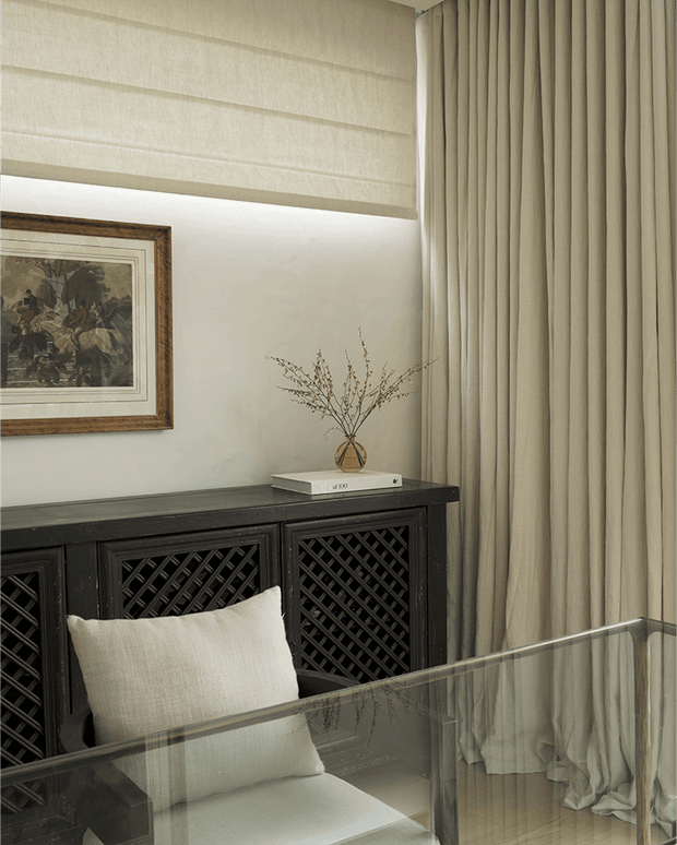 A refined, softly lit interior corner featuring a black carved lattice-front sideboard, a cream upholstered chair, layered beige curtains and Roman blind, a framed artwork, and a glass-topped table or desk. The palette is warm and neutral with traditional detailing and restrained contemporary styling.