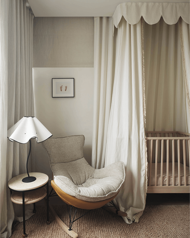 A softly lit nursery corner with a wooden cot screened by floor-to-ceiling cream curtains, a sculptural upholstered lounge chair, side table, floor lamp, textured carpet, and framed artwork. The palette is warm and neutral, with gently scalloped curtain detailing creating a cozy, refined atmosphere.