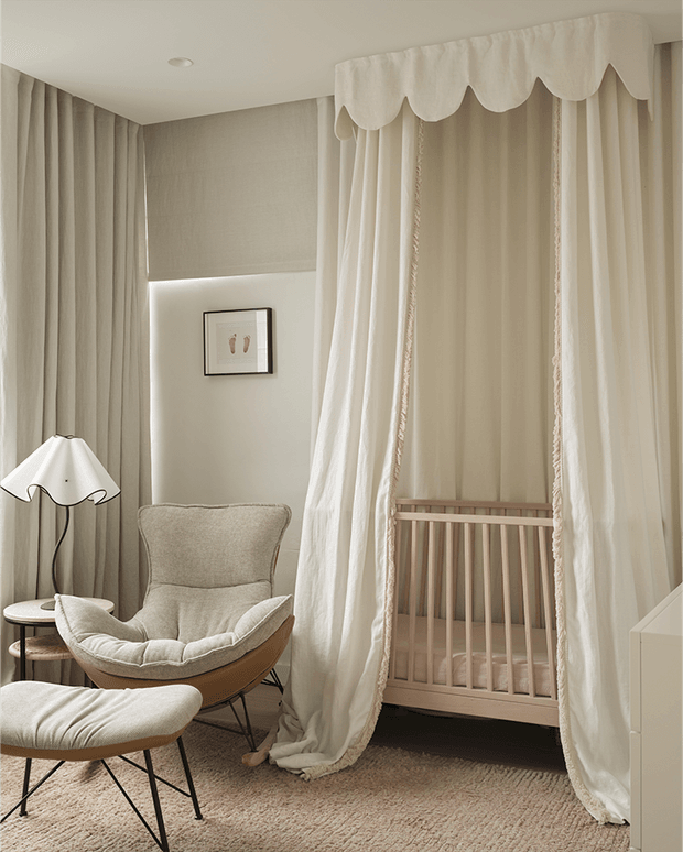 A softly styled nursery corner in warm neutral tones, featuring a wooden crib enclosed by a floor-to-ceiling cream canopy with a scalloped valance and draped curtains. The room includes a plush upholstered lounge chair and ottoman, a small round side table with a sculptural lamp, full-height curtains, textured carpet, and subtle wall artwork.