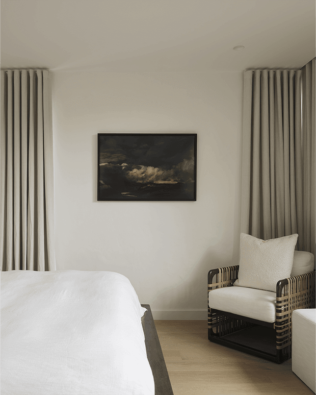 A calm, minimalist bedroom with a partially visible bed dressed in white linen, full-height neutral curtains framing the room, a black-framed dark landscape artwork, and a woven dark wood or rattan armchair with a cream cushion. Pale walls and light timber flooring create a warm, restrained interior.