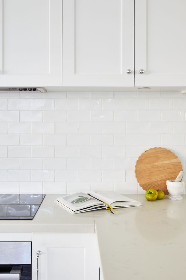 Bright, compact white kitchen with shaker-style upper and lower cabinetry, glossy white subway-tile splashback, pale stone benchtops, and a black glass cooktop. Open cookbooks, fruit, a white mug, and a round timber serving board sit on the benchtop, adding warm natural contrast to the predominantly white scheme.