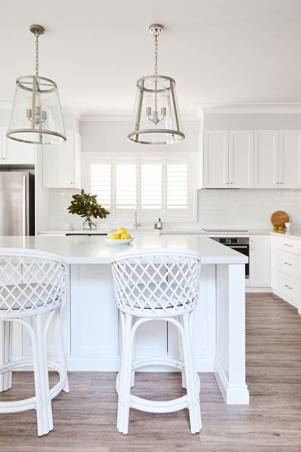Bright white kitchen with a large island and two white woven bar stools, white shaker-style cabinetry, a white subway-tile backsplash, pale timber-look flooring, stainless-steel appliances, and glass pendant lights. Plantation shutters and fresh styling create an airy coastal feel.