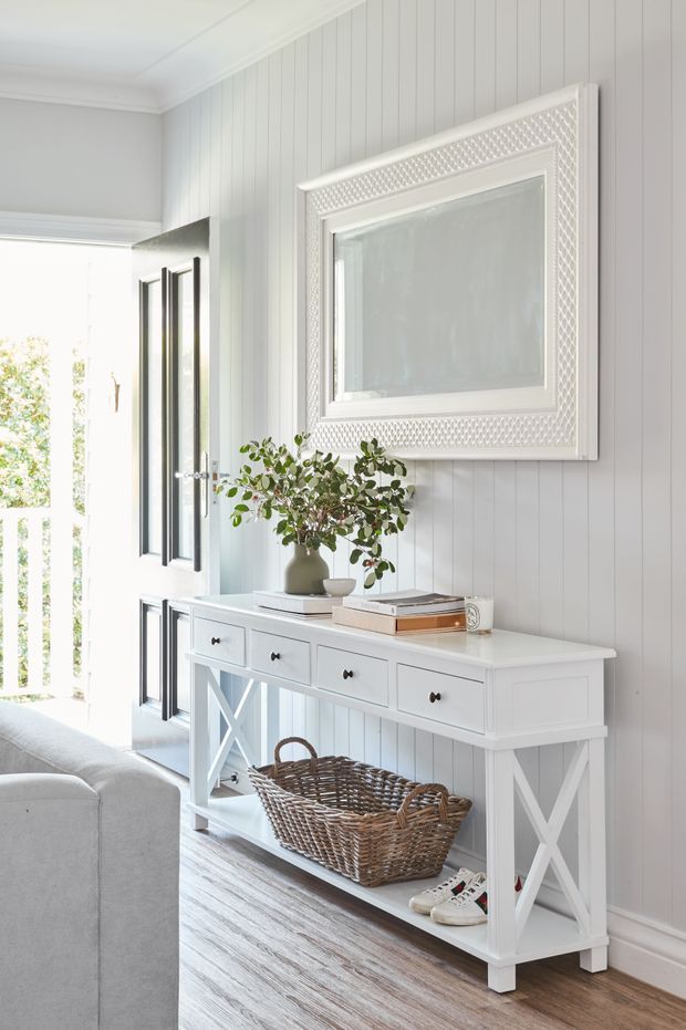 Bright, airy entry or hallway vignette featuring white vertical wall panelling, a white framed decorative mirror, a farmhouse-style console table with X-brace supports, woven basket storage, books, and a potted branch arrangement. An open glazed door provides strong natural light, while part of a light upholstered sofa is visible in the foreground. The styling is coastal and traditional with a clean, neutral palette.