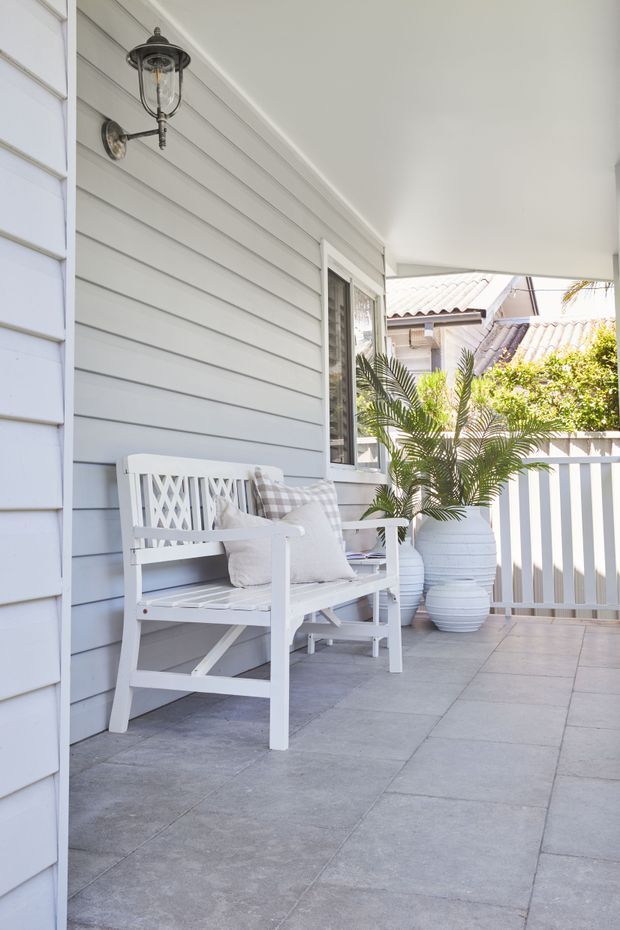A bright covered front porch or terrace with pale grey horizontal weatherboard cladding, white trim and balustrade, grey tiled flooring, a white lattice-back bench with cushions, and large white planters containing palm plants. A traditional lantern-style wall light and neighbouring tiled rooflines are visible, creating a relaxed coastal residential entry setting.