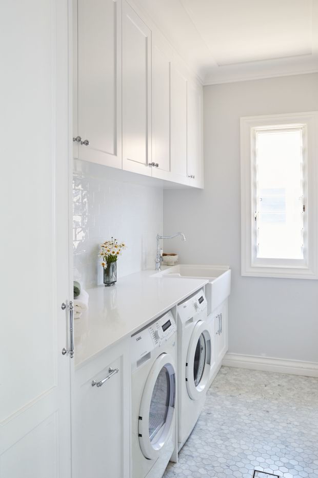 Bright, compact laundry room with white built-in cabinetry, a white countertop and integrated double sink, front-loading washer and dryer, glossy white tiled splashback, and a pale hexagonal floor tile. The room has soft grey walls, white decorative trim, chrome hardware, and a tall window providing abundant natural light.