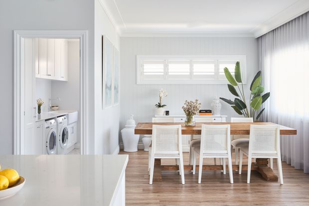 Bright open-plan dining area viewed from a kitchen, with a long natural timber dining table, white woven-back chairs, pale grey walls, white trim, timber-look flooring, sheer curtains, and a large tropical indoor plant. A compact white laundry with washer and dryer is visible through a doorway on the left. The scene has a clean, airy, contemporary coastal feel.