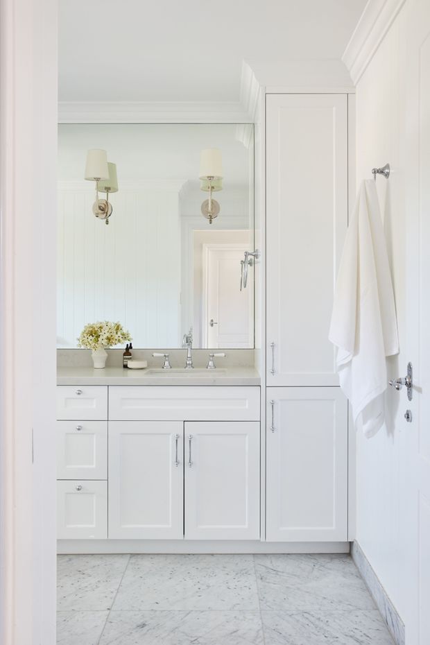 Bright, refined bathroom vanity area with white paneled cabinetry, a pale stone countertop, polished chrome fixtures, large mirrored wall, wall sconces, and light veined marble floor tiles. A tall built-in storage cabinet and white towels occupy the right side, while the room is finished with traditional moulding and white vertical wall panelling.