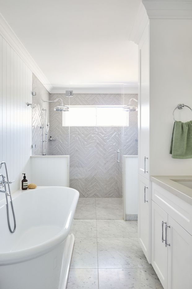 Bright, elegant white bathroom featuring a freestanding bathtub, glass-enclosed shower, double shower fixtures, white vanity cabinetry, marble-look tiled flooring, and a herringbone-patterned grey wall tile. The room has traditional detailing including wall panelling and decorative crown moulding, with chrome hardware and soft natural light.
