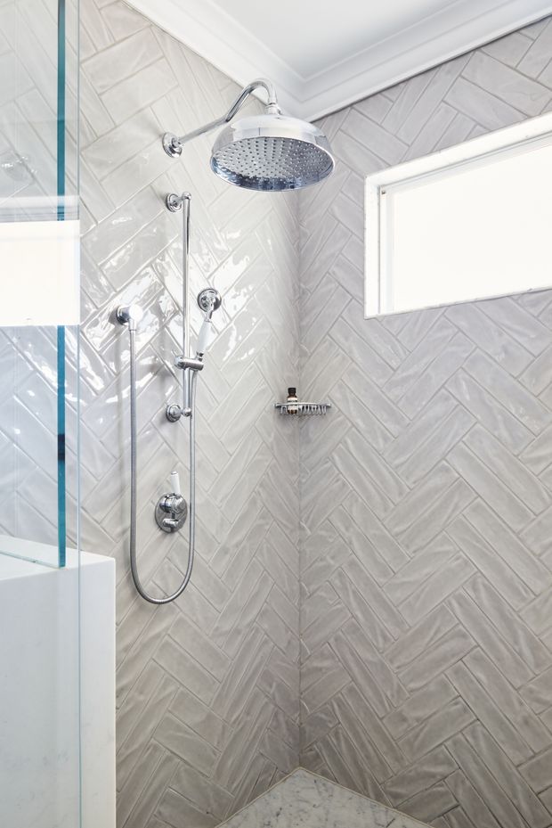 Bright, close-up view of a contemporary walk-in shower enclosure with glossy light-grey herringbone wall tiles, chrome rainfall showerhead and handheld shower fittings, a small built-in shelf, frameless glass screen, white trim, and a small frosted window.