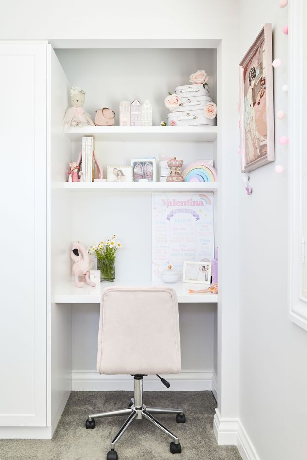 Bright, compact built-in study nook set into a wall alcove, with white shelving, a pale upholstered swivel office chair, and softly styled children's keepsakes, books, framed photos, toys, and pastel decorative objects. White trim and cabinetry surround the niche, while grey carpet covers the floor. A framed artwork and pink-and-white decorative light garland add a playful touch.