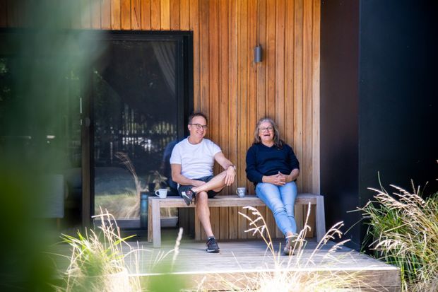Exterior view of a contemporary dark-clad dwelling with a warm vertical timber wall and large glazed doorway. Two people sit on a simple built-in or freestanding bench on a timber deck, surrounded by ornamental grasses and garden planting.