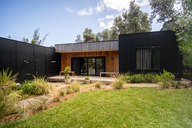 Single-storey contemporary house with a low, dark-clad facade and a recessed central entry or living zone. Black exterior cladding contrasts with warm vertical timber cladding around the glazed doors, while a small timber deck opens onto a landscaped garden with ornamental grasses and established trees.
