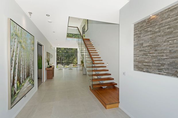 Bright contemporary residential hallway and stairway with a floating timber staircase enclosed by clear glass balustrades. The space features large pale tiled flooring, white walls, recessed ceiling lighting, a double-height stair void, a large framed artwork, and a textured grey stone feature wall.
