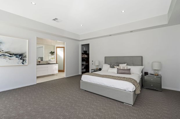 A spacious, contemporary master bedroom with a large upholstered grey bed and matching bedside tables, layered white and patterned bedding, and wall-to-wall grey carpet. The room has recessed ceiling lighting, light grey walls, minimalist styling, built-in shelving, and an open connection to a bright ensuite bathroom with a double vanity. Abstract artwork adds a subtle accent.