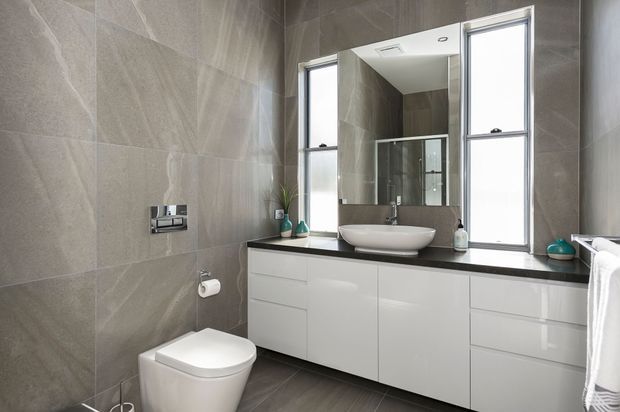 Contemporary bathroom with large grey stone-look wall and floor tiles, a wall-mounted dual-flush toilet, glossy white floating vanity cabinetry, dark benchtop, vessel basin, large mirror, and tall windows providing natural light.