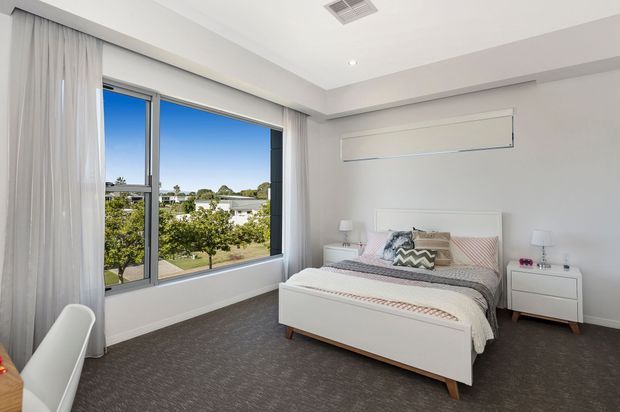 Bright contemporary bedroom with a white upholstered bed, matching white bedside tables, soft grey and blush bedding, floor-to-ceiling curtains, a large corner window, and dark grey carpet. The room overlooks a leafy suburban neighbourhood and has a clean, restrained finish with recessed ceiling lighting and concealed wall-mounted air conditioning.