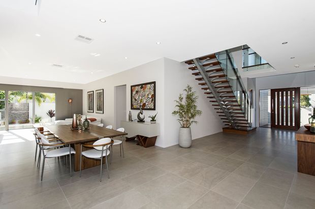 Bright, open-plan contemporary entry and dining area with a large timber dining table, upholstered chairs, white walls, large grey floor tiles, framed artwork, indoor plants, and a floating timber-and-metal staircase with glass balustrade. A glazed front door and outdoor landscaping are visible beyond the entry.