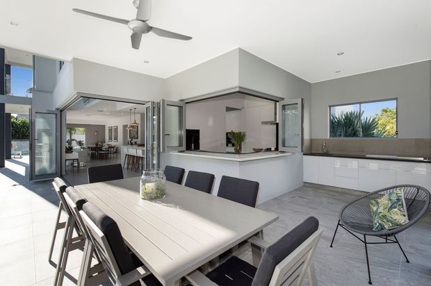 Bright, open-plan contemporary kitchen and dining area with a large light-toned dining table, dark upholstered chairs, white island cabinetry, grey tiled flooring, and extensive folding or sliding glass doors opening to an outdoor area. A ceiling fan, recessed lighting, expansive windows, and a partially visible living space contribute to the airy indoor-outdoor layout.