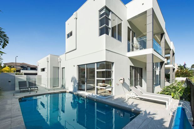 A bright contemporary multi-storey white rendered residence viewed from the rear courtyard, featuring large glazed openings, projecting balconies with glass balustrades, a paved pool terrace, and a rectangular swimming pool. The scene has strong geometric forms, clean lines, and abundant sunlight.