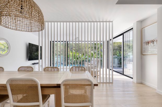 Bright contemporary dining area with a pale timber dining table and woven cane chairs. A large woven pendant light hangs above the table, while a vertical white timber-screen partition separates the dining space from a living area and opens toward floor-to-ceiling glazing and a garden beyond. The room has white walls, pale timber flooring, black-framed glazing, and restrained coastal-natural styling.