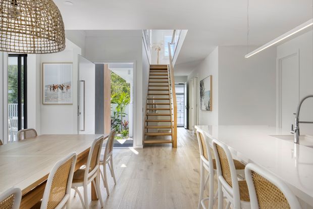 Bright open-plan interior combining a dining area and white kitchen, with pale timber flooring, woven rattan dining chairs, a large woven pendant light, and a central open-tread timber staircase. White walls, cabinetry and linear lighting create a clean contemporary coastal feel, while an open doorway leads to a planted outdoor area.