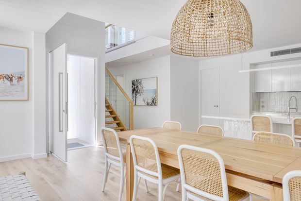 Bright open-plan dining and kitchen area with a large light timber dining table, white-framed cane dining chairs, woven pendant light, pale timber flooring, and white cabinetry. A glazed entry door and timber staircase lead toward an upper level, while the kitchen is visible at right beneath a high ceiling and mezzanine-like opening.