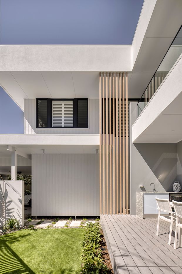 A bright contemporary multi-level residential courtyard viewed from an outdoor deck. White rendered walls and soffits frame open balconies and a central vertical timber batten screen. Black-framed louvred windows, glass balustrades, and a compact outdoor kitchen with pale stone benchtop sit beside a grey timber-look deck. Artificial lawn, stepping stones, and planting occupy the courtyard below.