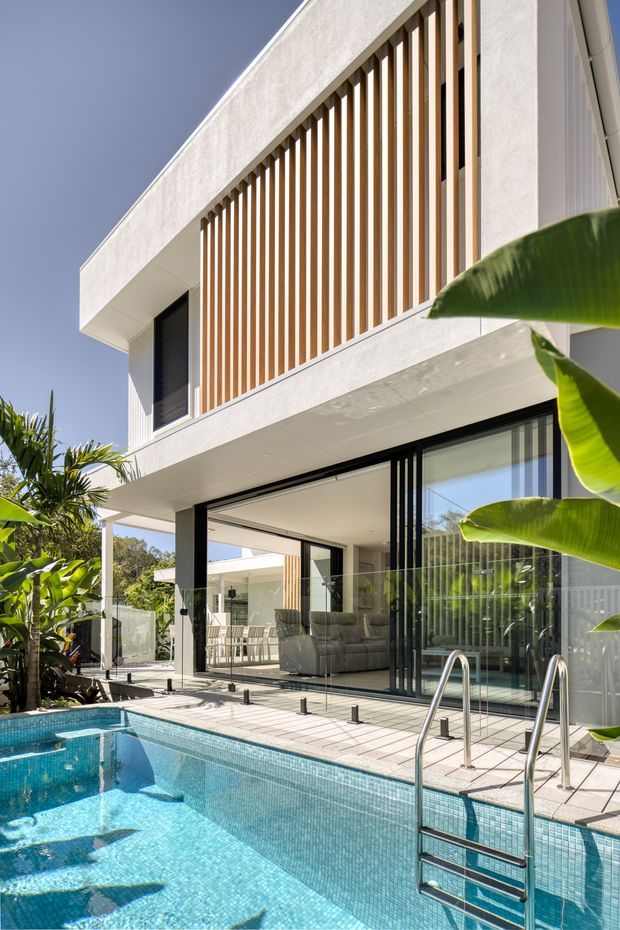 Bright exterior view of a contemporary two-storey residence with white rendered walls, extensive black-framed glazing, and a prominent upper-level screen of warm timber-look vertical battens. The open ground floor connects to a tiled pool terrace, with a clear blue swimming pool, stainless-steel ladder, glass balustrade, and tropical planting around the edges. The living area and outdoor dining zone are visible through the open glazing.