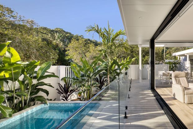 A bright contemporary outdoor living area beside a narrow swimming pool, with a timber-look deck, frameless glass balustrade, lush tropical planting, and a covered pavilion. Large sliding glass openings connect the deck to a furnished lounge and dining area, while white walls and slim dark structural frames create a clean resort-like setting.