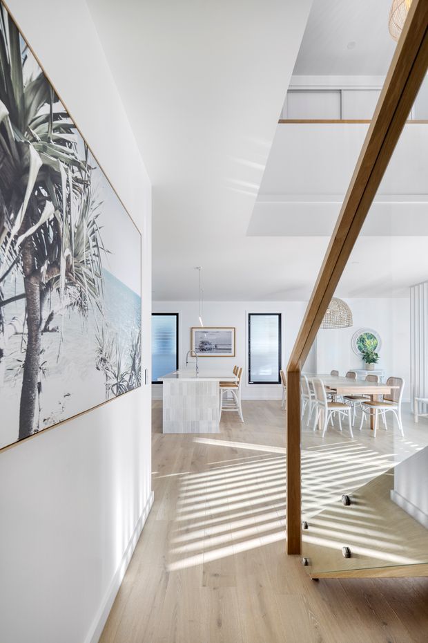 Bright contemporary open-plan interior viewed from a stair landing or hallway, looking toward a white kitchen and dining area. The space features pale timber flooring, white walls and ceilings, a prominent timber stair handrail and structural post, large framed tropical artwork, woven pendant lights, a white island, and a light timber dining table with white chairs. Strong sunlight creates striped shadows across the floor, while a double-height void and upper-level balustrade add vertical openness.