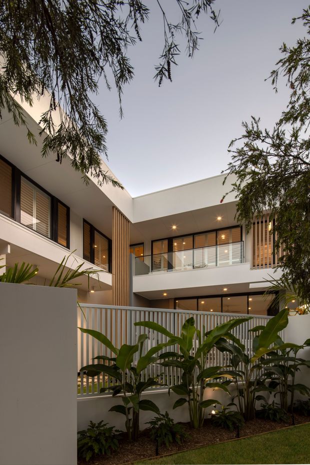 A contemporary multi-level residential courtyard viewed from a landscaped garden. The building has clean white rendered volumes, broad flat rooflines, dark-framed glazing with external shutters, warm timber-toned vertical screening, recessed balcony spaces, and illuminated soffits. Tropical planting and a slatted privacy fence occupy the foreground, creating a lush enclosed outdoor setting.