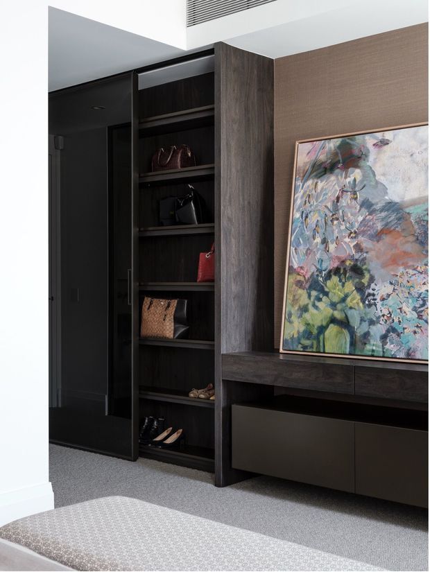A refined entryway or dressing area with dark timber built-in shelving displaying handbags and shoes, integrated low cabinetry, and a large framed abstract artwork. The space has charcoal and deep wood tones, textured taupe wall covering, grey carpet, and crisp white architectural surfaces, creating a moody, sophisticated interior.