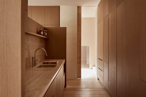 A narrow, refined galley kitchen viewed toward a bright adjoining hallway. Warm timber cabinetry lines both sides, with a stone benchtop and integrated sink on the left, discreet shelving and upper cupboards, and full-height built-in storage on the right. Soft natural light enters from the rear, creating a calm, minimalist interior with strong linear joinery.