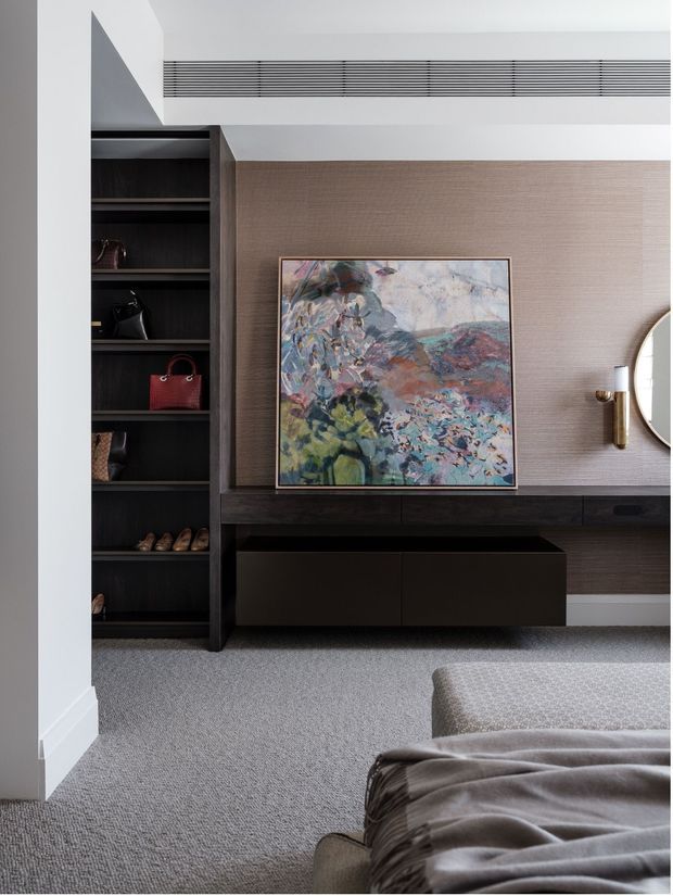 A refined bedroom interior featuring dark timber built-in shelving and a low media or storage console, a textured taupe feature wall, abstract artwork, brass wall sconce, partial round mirror, and soft grey carpeting. The edge of a bed and upholstered bench are visible in the foreground. The room has clean contemporary joinery and a muted, sophisticated palette.