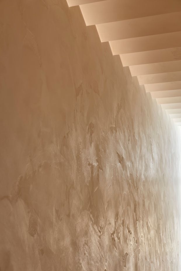 Close-up view of a warm beige textured plaster wall beneath a series of evenly spaced white ceiling beams or rafters. The wall has a hand-applied, irregular tactile finish, while bright natural light enters from the far end of the narrow space. The image focuses on the architectural surface and ceiling rhythm rather than a furnished room.