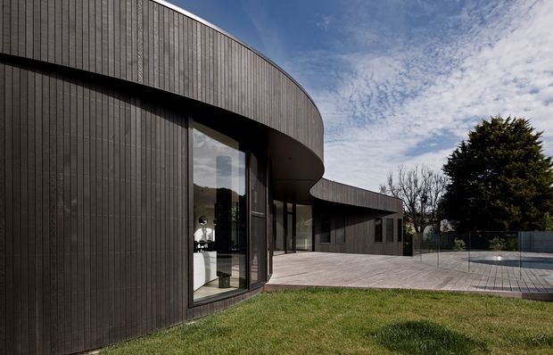 Exterior view of a contemporary, low-profile building with a dramatic curved facade and projecting roofline clad in dark vertical timber boards. Full-height glazing opens toward a broad timber deck and paved outdoor area, with a simple landscaped lawn and mature trees beyond.