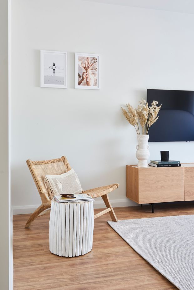 A bright, softly styled living room with pale walls, warm timber flooring, a woven rattan lounge chair, a sculptural white side table, and a low timber media console beneath a wall-mounted television. Neutral artwork, dried grasses, and a light rug create a calm, contemporary natural aesthetic.