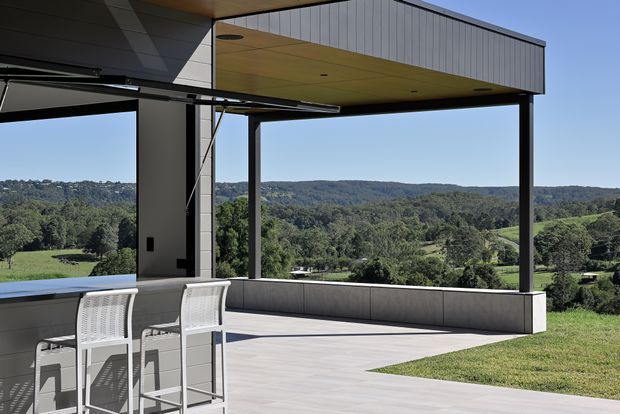 Contemporary covered outdoor terrace overlooking a green, rolling landscape. The space features a slim dark structural frame, a projecting flat roof with a pale vertically panelled fascia and recessed ceiling lights, expansive open sides, a built-in concrete-toned perimeter bench or planter wall, and a light paved floor. A bar-height outdoor table and chairs sit in the foreground, with a partially visible pool or water feature at left.