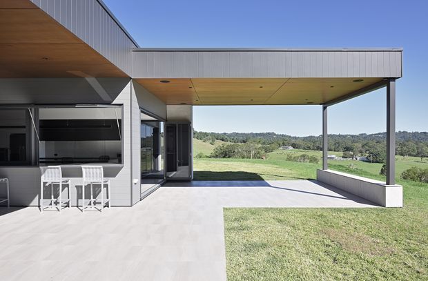 A modern residential outdoor entertaining area with a broad flat-roofed covered terrace extending over a concrete patio. The open-sided pavilion features slim dark structural posts, a warm timber-lined soffit, pale metal wall cladding, and a built-in outdoor kitchen with bar seating at the left. The terrace overlooks a rural green landscape and rolling hills.