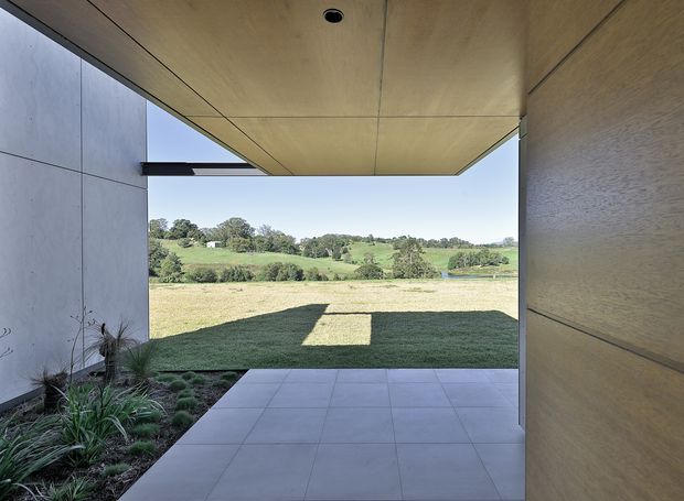 A covered outdoor passage or entry terrace framed by smooth light-coloured wall panels and a projecting timber-lined ceiling. Large square paving tiles lead toward an open rural landscape with rolling green fields and scattered trees. The space has a strong indoor-outdoor connection, deep overhang, simple modern detailing, and landscaped planting along one edge.