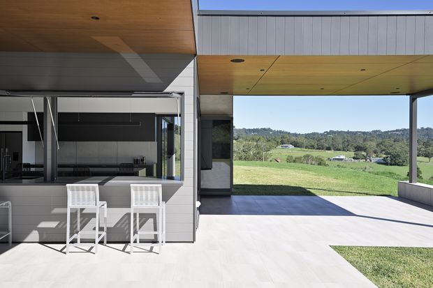 A contemporary covered outdoor living area beside a compact outdoor kitchen or bar, framed by a light grey paneled facade and warm timber-lined soffits. White bar stools sit at the built-in counter, while large open glazing and the broad roofed terrace overlook a green rural landscape. The space is bright, open, and strongly connected to the outdoors.