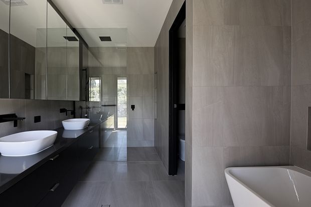 A contemporary bathroom with a long double vanity in dark cabinetry, two white vessel basins, wall-mounted black tapware, extensive mirrored cabinetry, large-format grey stone-look wall and floor tiles, and a freestanding white bathtub in the foreground. A glazed shower area and bright exterior view are visible at the rear.