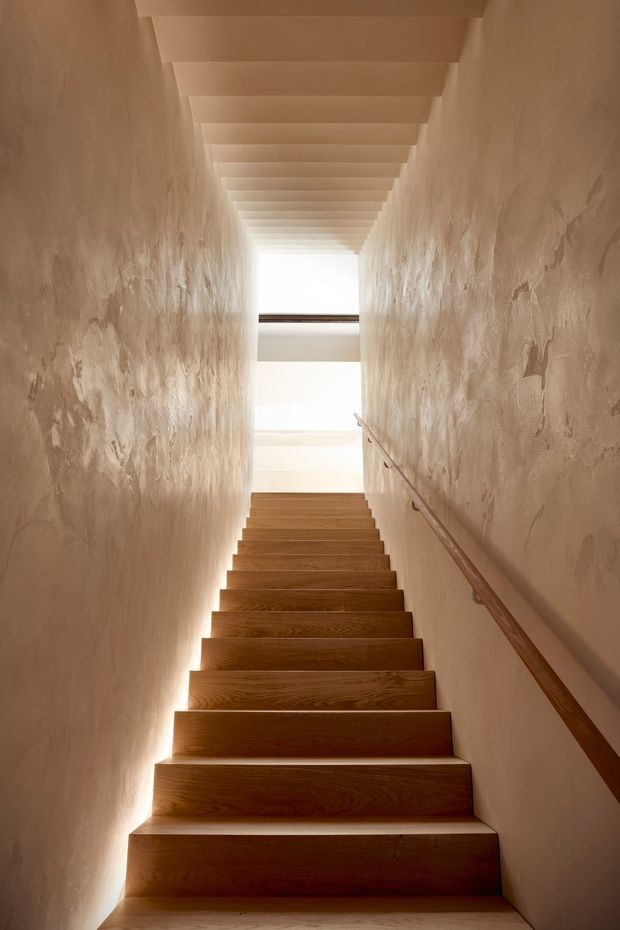 A narrow, tall stairway viewed from the bottom, with warm timber stairs and a timber handrail running along one side. Rough textured plaster walls enclose the stairwell, while a series of exposed white-painted ceiling beams or rafters leads toward a bright upper landing, creating a strong linear perspective and softly illuminated, warm atmosphere.