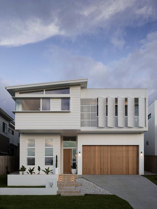 Front exterior view of a contemporary two-storey residence with white horizontal cladding, a broad low-pitched roof, projecting upper-level glazing, and a prominent white vertical screening system over the right-side upper facade. The ground floor includes a timber garage door, recessed entry, concrete driveway, gravel landscaping, and a low white rendered boundary wall.