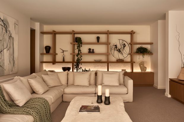 Warm, refined living room with a large cream L-shaped sectional sofa, textured throw, soft upholstered coffee table, and low-profile timber cabinetry. A prominent open timber display shelving system spans the rear wall, holding curated ceramics, sculptures, books, and trailing greenery. Ambient concealed lighting, neutral plaster walls, carpeted flooring, and abstract artwork create a calm contemporary atmosphere.