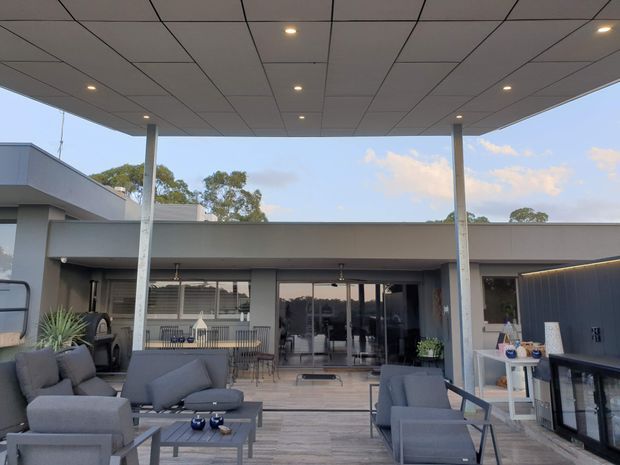 A covered outdoor entertaining area beside a modern single-storey home, furnished with grey lounge chairs and sofas around a low coffee table. The space has timber-look decking, a broad flat canopy with recessed lighting, slim support columns, glass sliding doors, and an outdoor kitchen or bar with dark vertical cladding on the right. A dining area and interior spaces are visible beyond the glazing.
