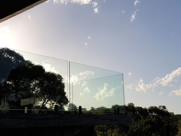 Exterior architectural view of a contemporary elevated terrace or balcony with frameless glass balustrades, silhouetted trees, and a broad sky beyond. The image is backlit by low sunlight, emphasizing the transparent glazing and projecting horizontal structure.